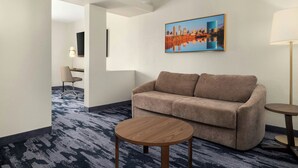 Une chambre d’hôtel avec un canapé, une table ronde en bois, une lampe, un tableau représentant un paysage urbain et un sol recouvert d’une moquette.