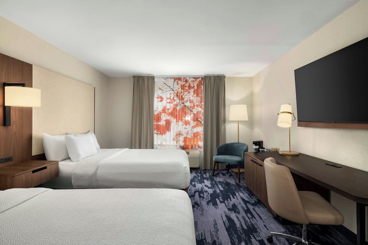 Ein Hotelzimmer mit zwei Betten, einem Schreibtisch, einem Stuhl, einem Fernseher und einem Fenster mit Vorhängen.