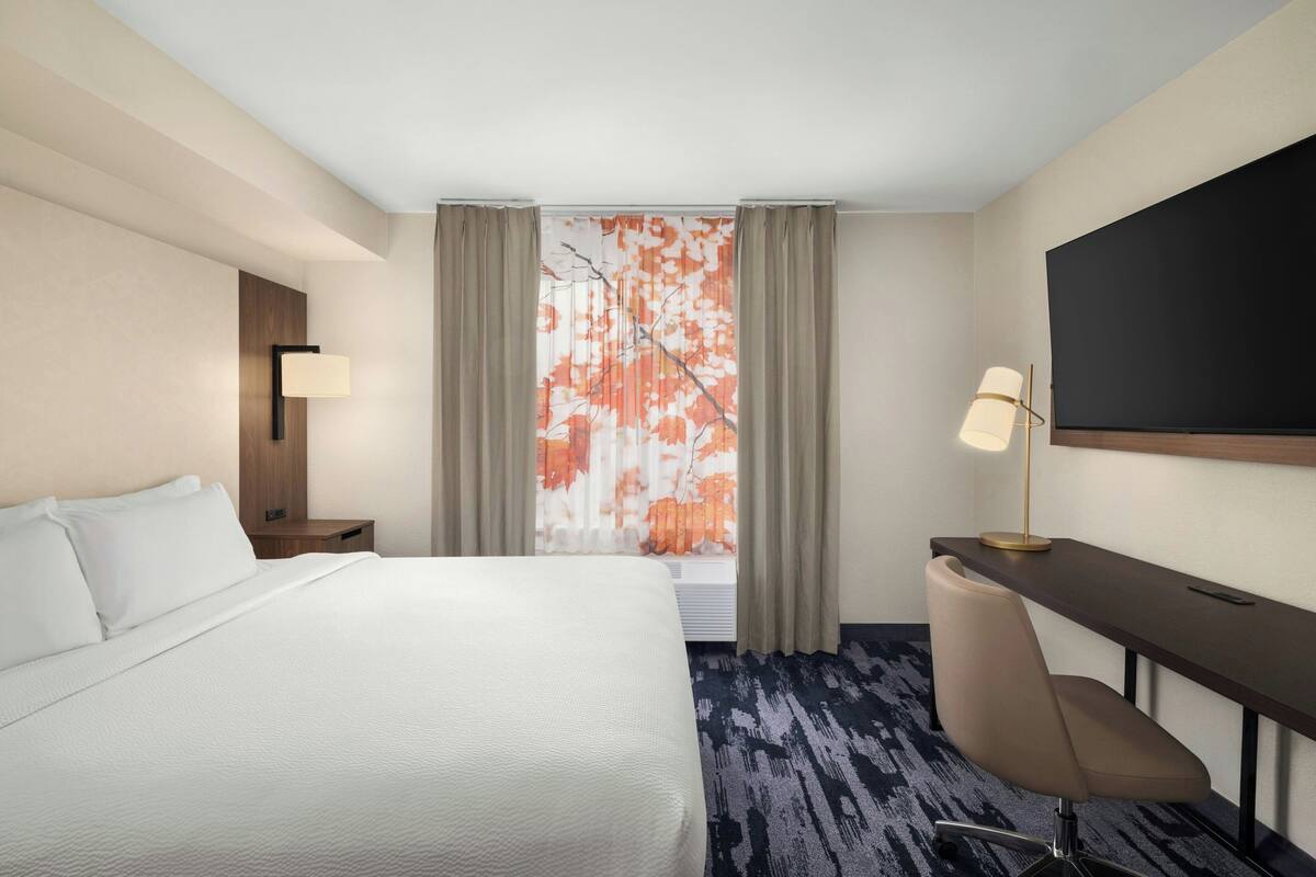 Ein Hotelzimmer mit Bett, Schreibtisch, Stuhl, Fernseher und einem Fenster mit Vorhängen.