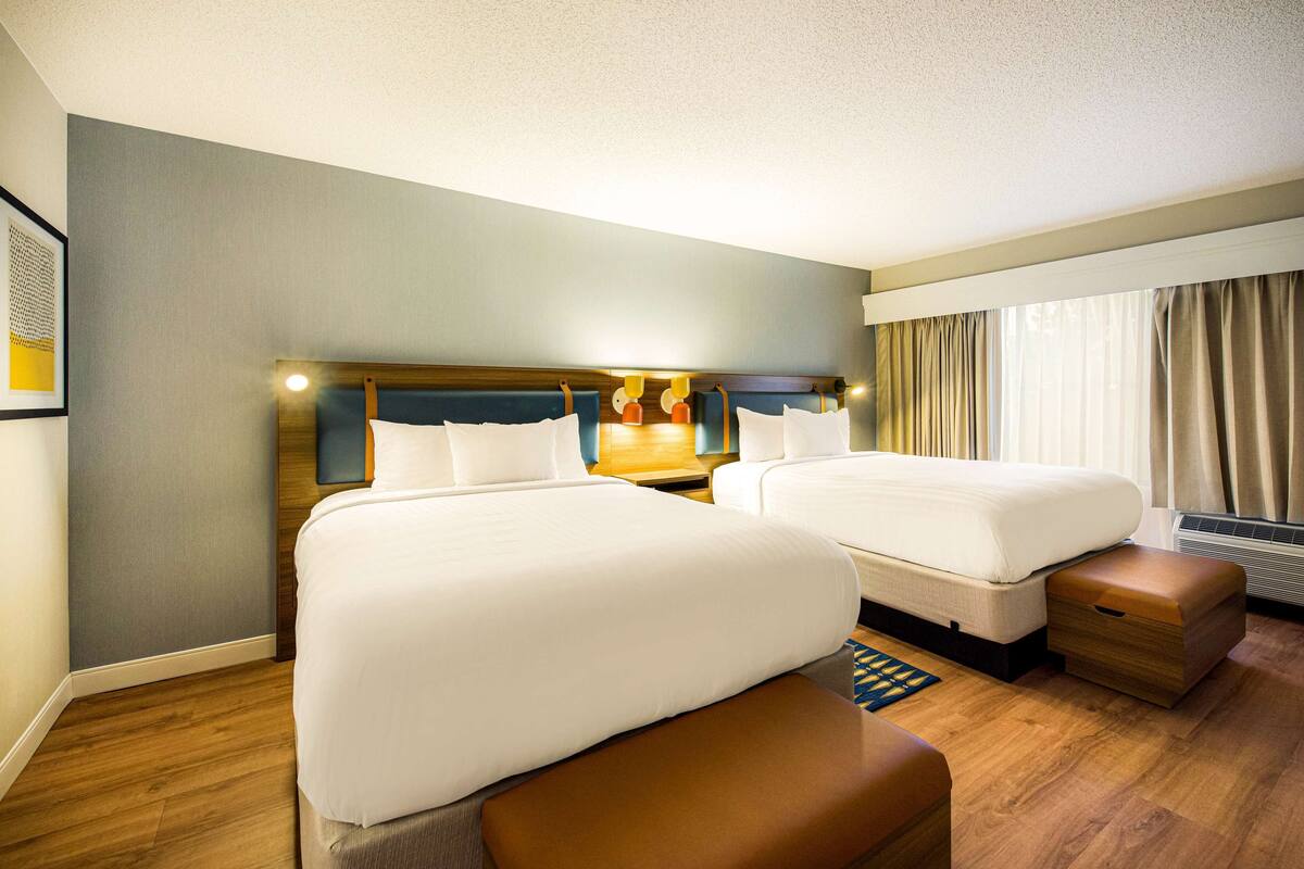 A hotel room with two beds, a wooden headboard, a nightstand, a bedside table, and a window with curtains.