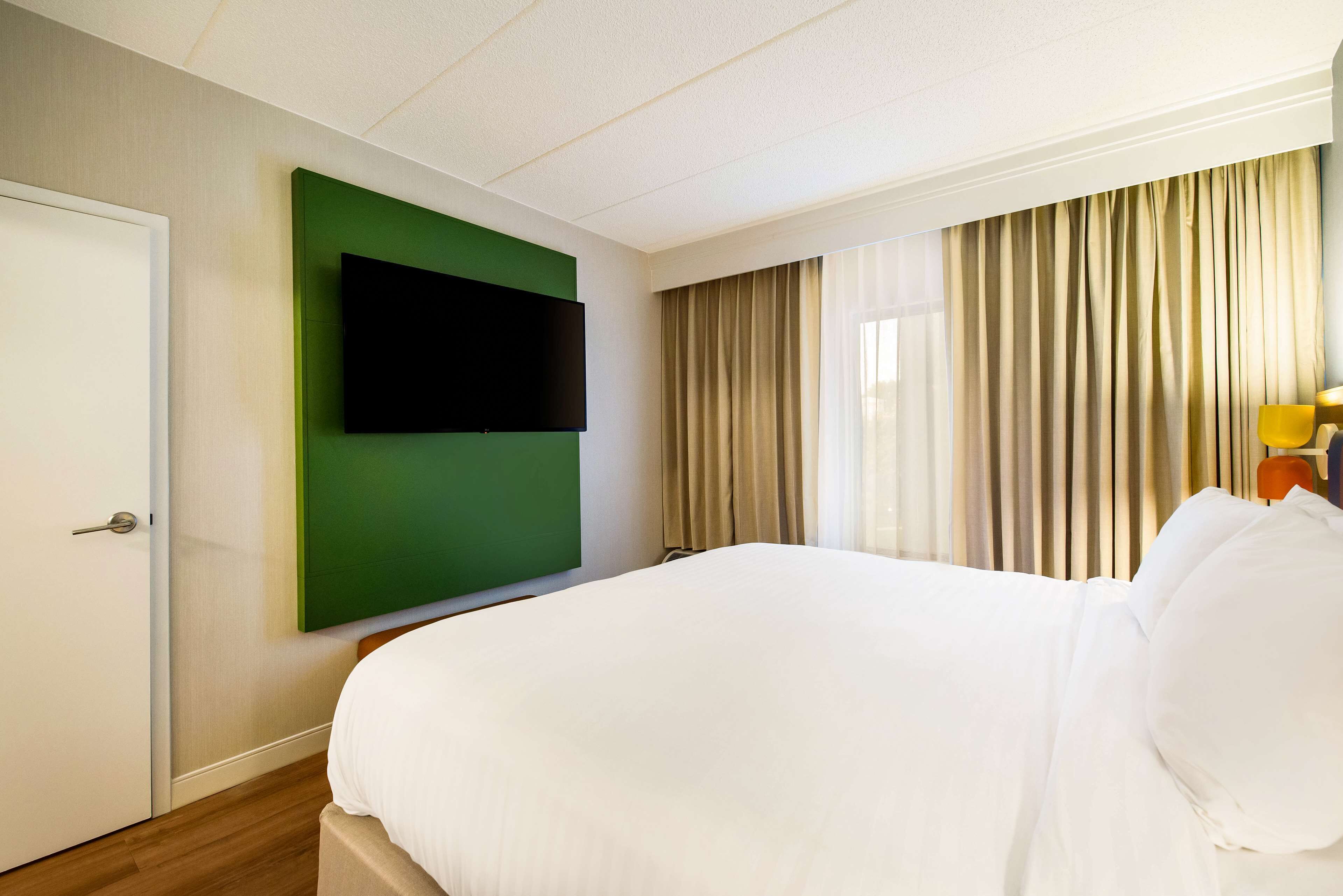 Une chambre d’hôtel avec un grand lit, un téléviseur à écran plat fixé sur un panneau vert, des rideaux beiges et un plancher en bois.
