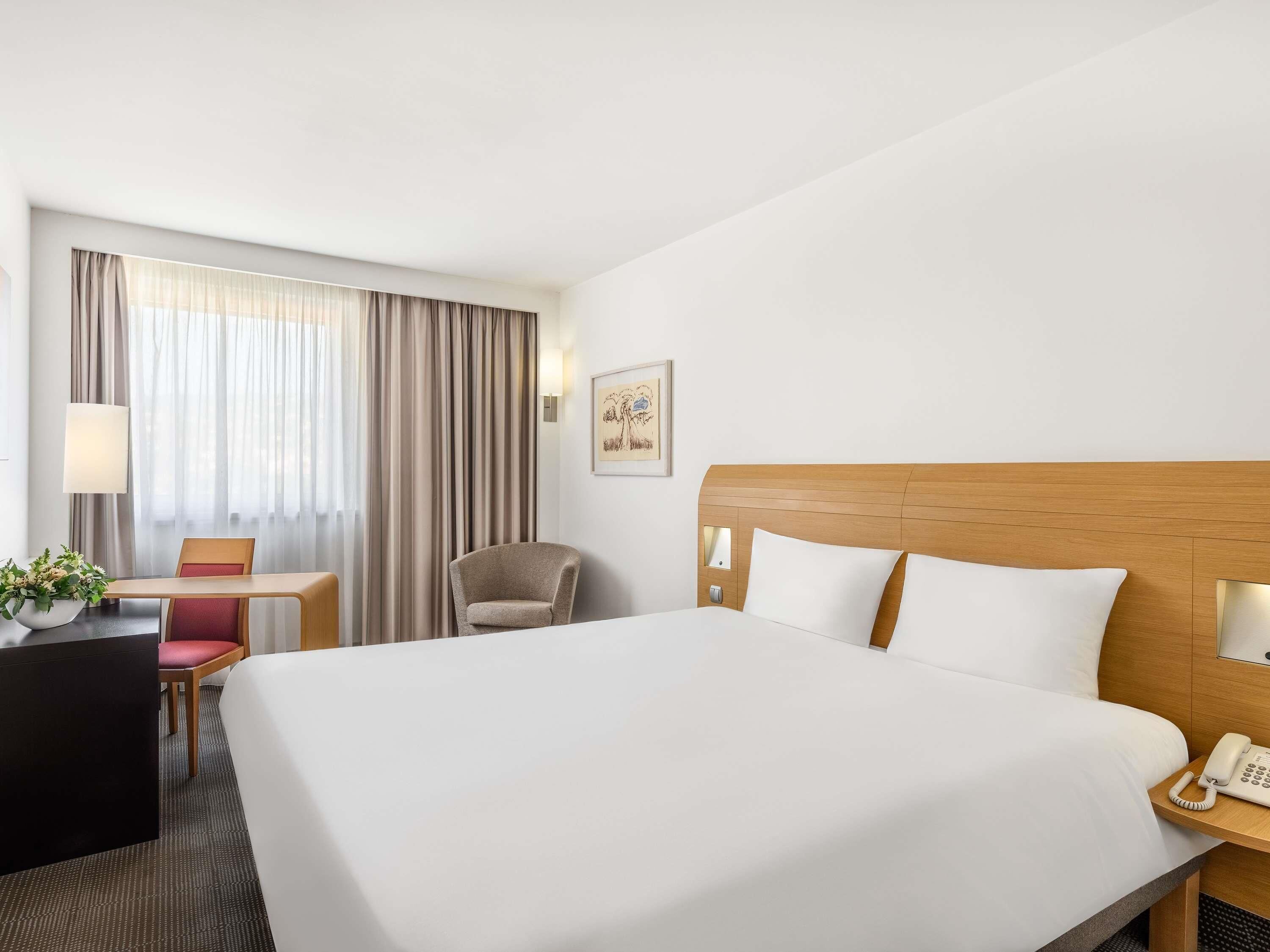 A hotel room with a large bed, a desk with a chair, a telephone, and a window with curtains.