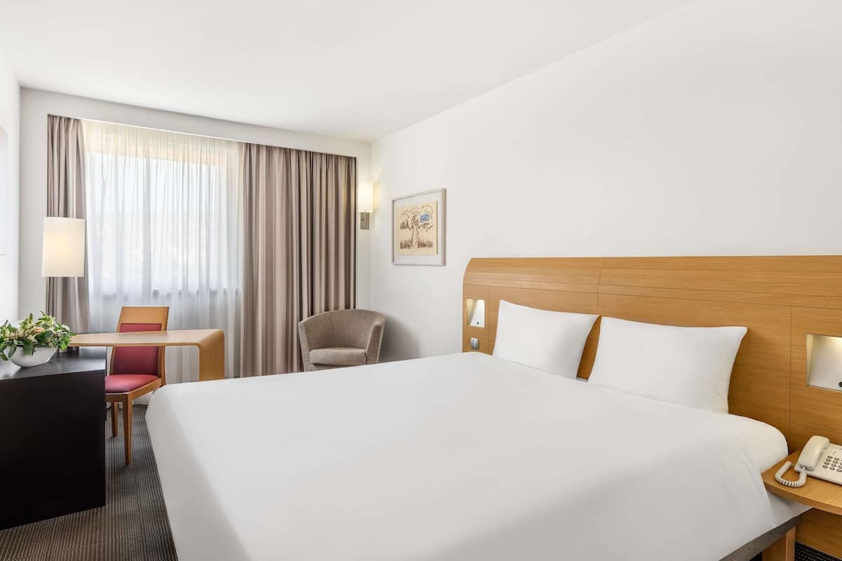 A hotel room with a large bed, a desk with a chair, a telephone, and a window with curtains.
