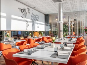 A modern restaurant with white tables and orange chairs, a world map on the wall, and a large pillar in the center. - Novotel Budapest City (Budapest)