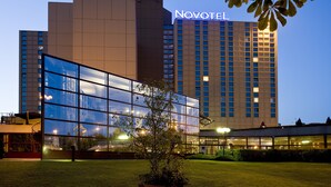 A modern hotel building with a prominent 'NOVOTEL' sign, large glass windows, and a well-maintained lawn area.