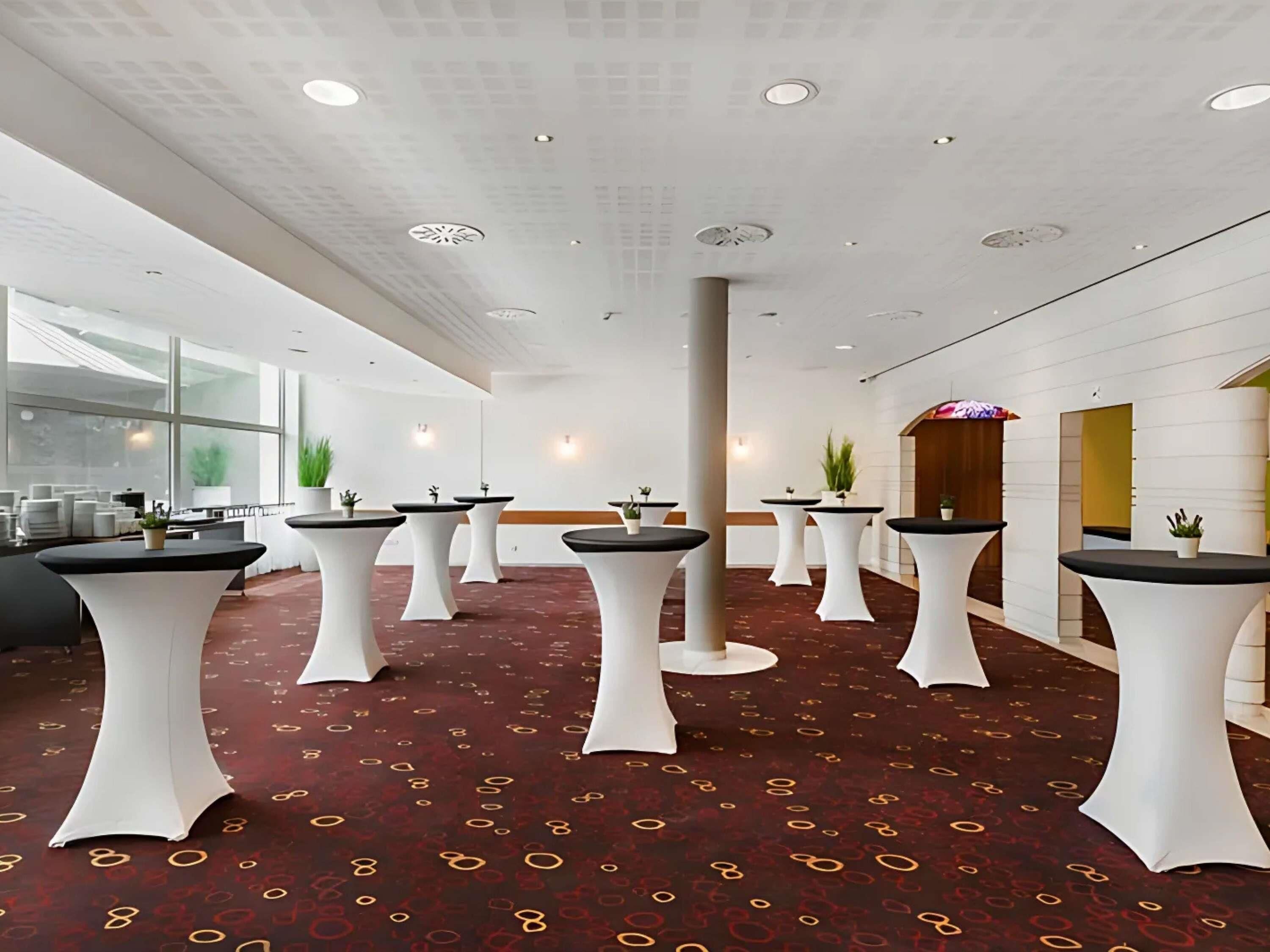 A modern hotel lobby with round tables, potted plants, and a patterned carpet.