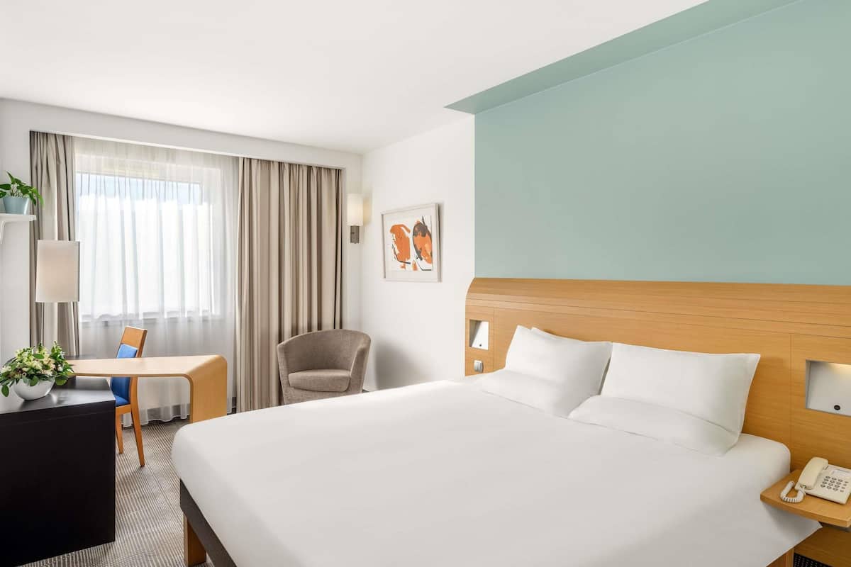 A hotel room with a large bed, a desk with a chair, a telephone, and a wall-mounted lamp.