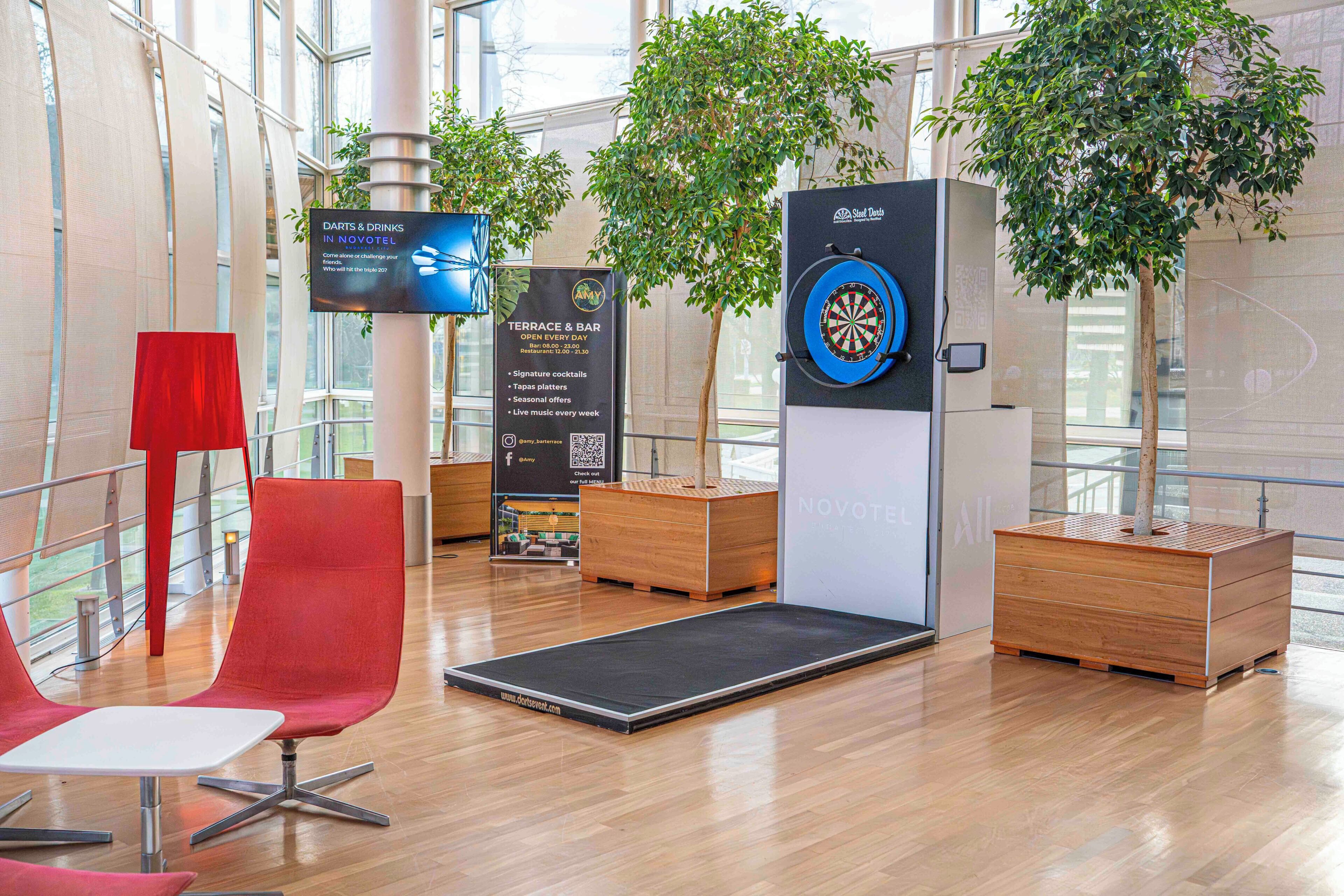 A modern hotel lobby with a dartboard, seating area, and a sign for the terrace and bar.