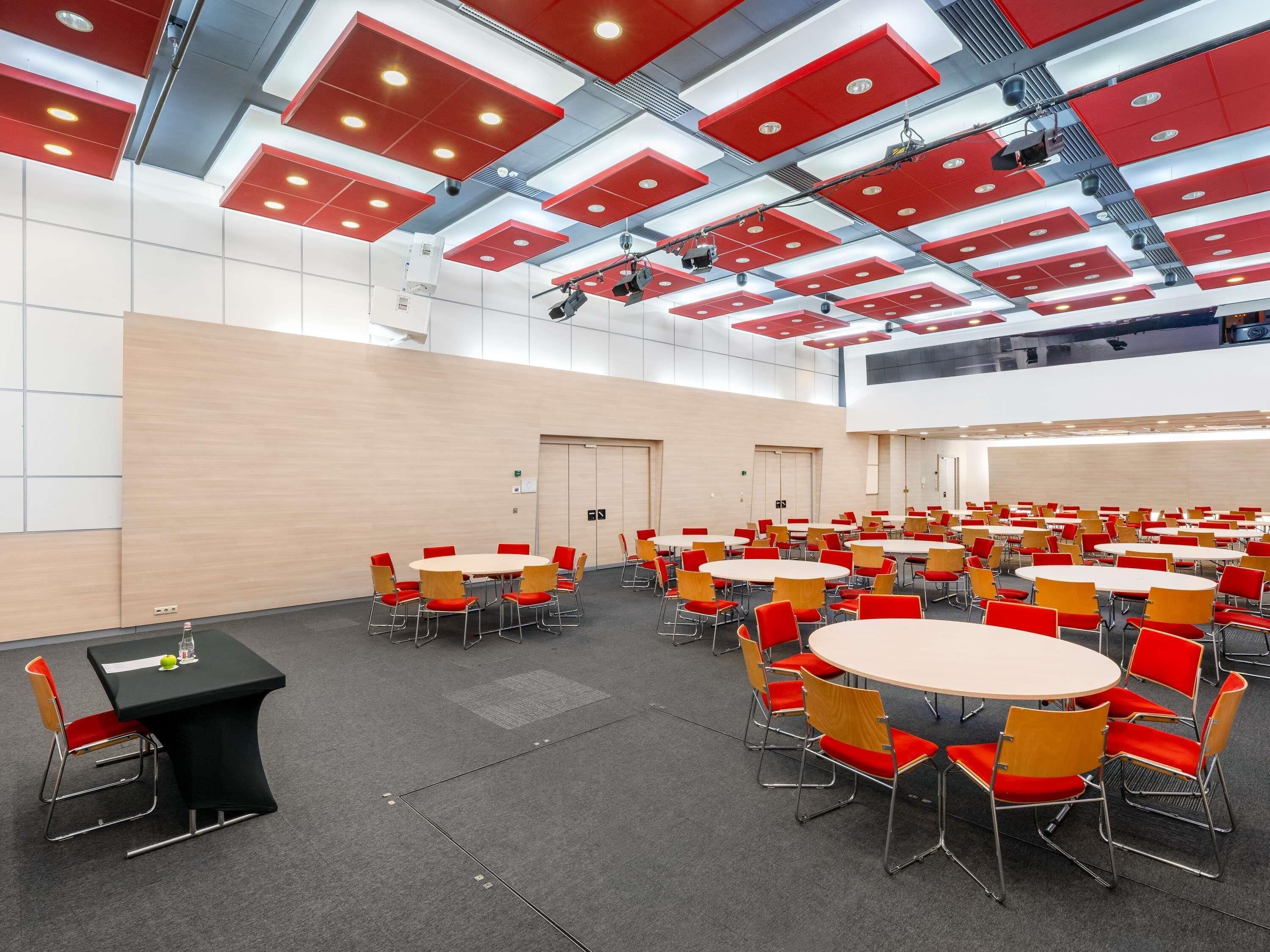Un salón de conferencias con varias mesas y sillas, un escenario con equipo de iluminación y un techo alto con paneles rojos.
