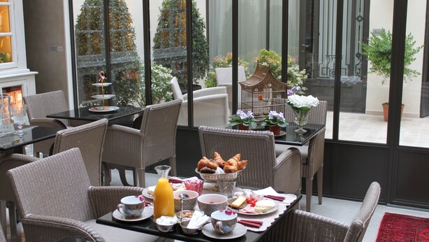 A dining area with wicker furniture, a table set with breakfast items, and a view of a garden through large windows. - My Home in Paris Hotel (Paris)