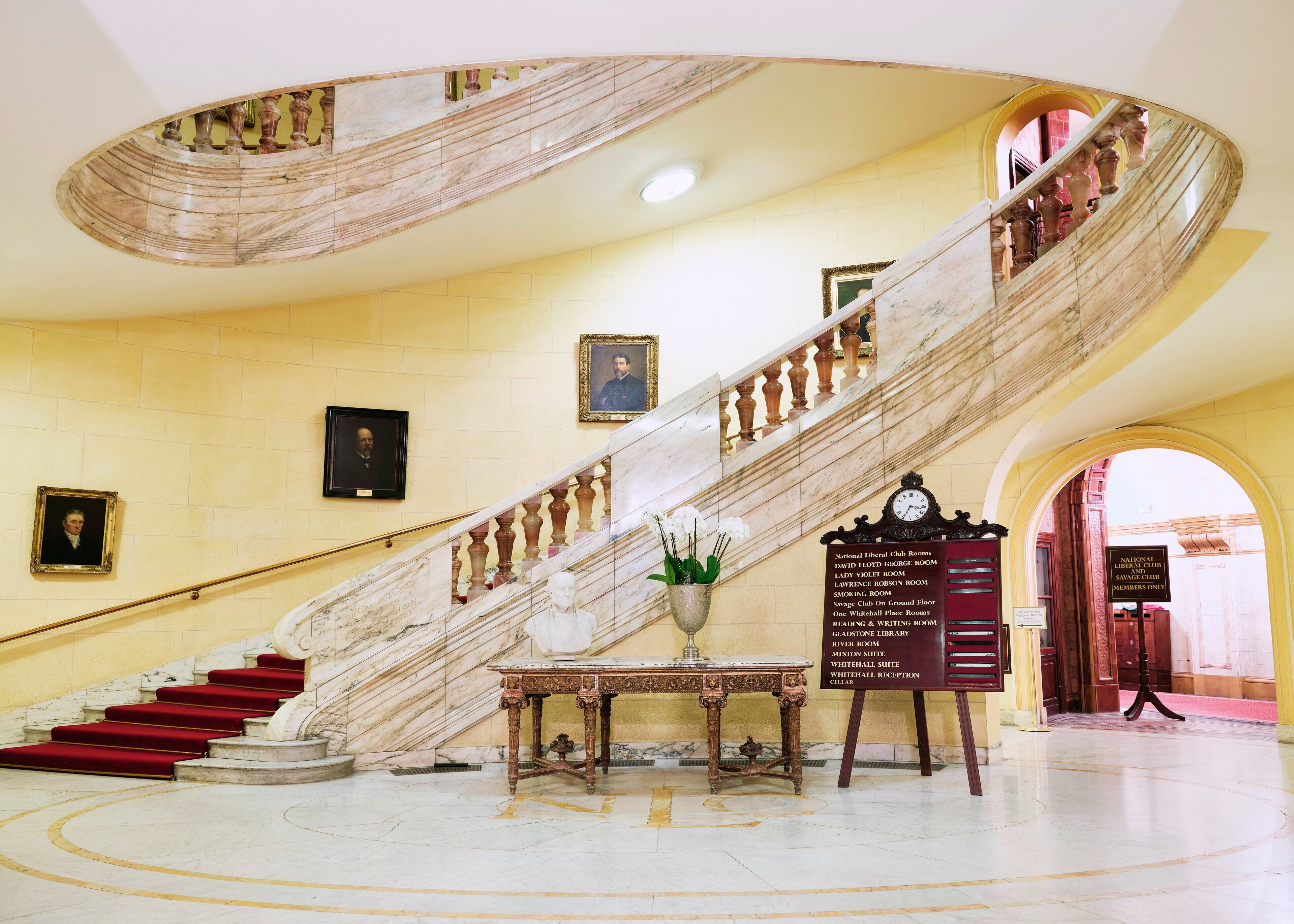 A grand marble staircase with red carpeted steps leading to a landing with a table and a sign listing room names.