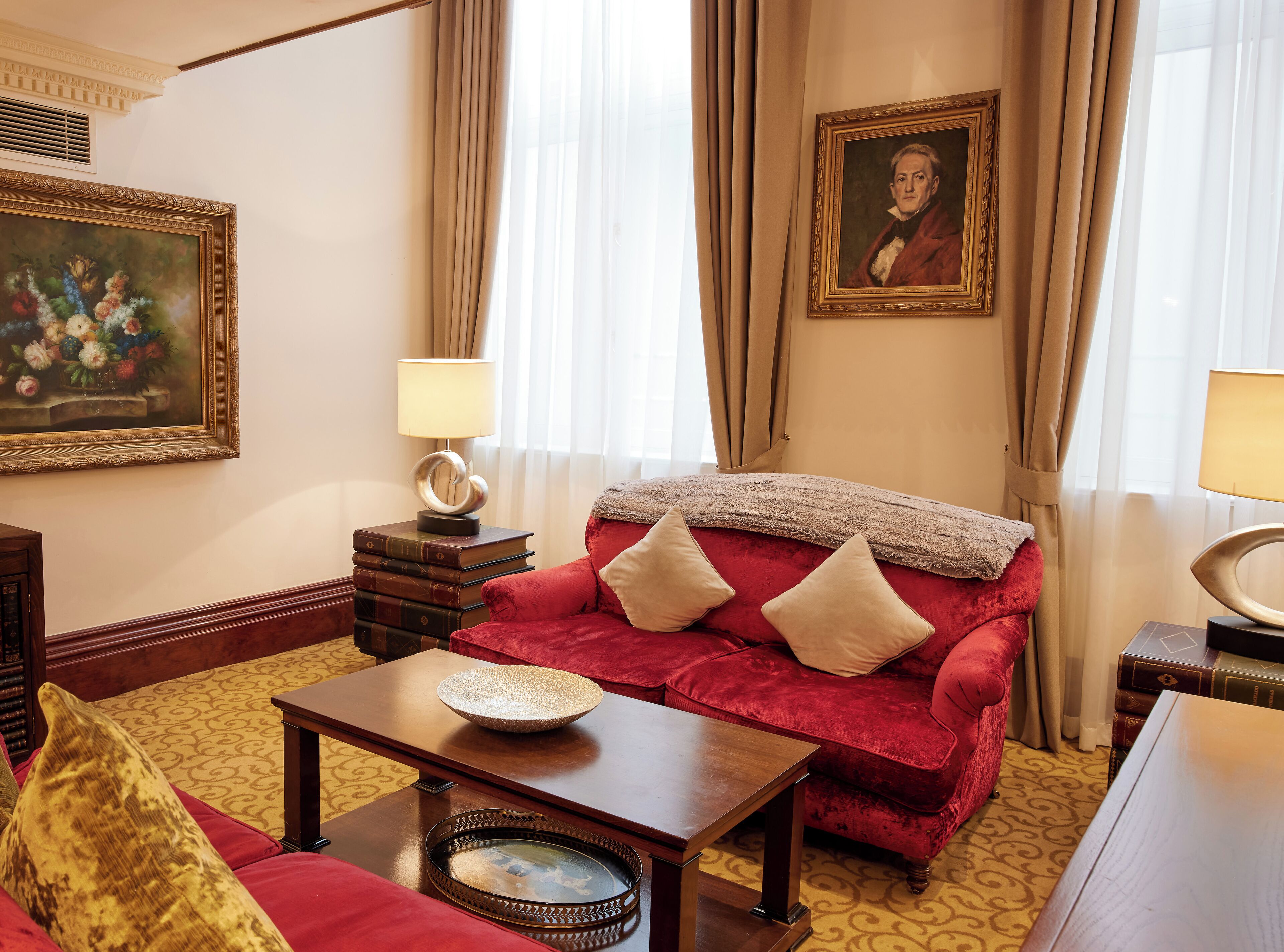 Um quarto com um sofá de veludo vermelho, uma mesa de centro de madeira, um tapete estampado, um quadro emoldurado e um abajur sobre uma mesinha.