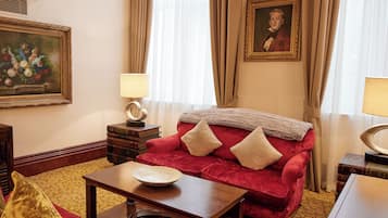 Um quarto com um sofá de veludo vermelho, uma mesa de centro de madeira, um tapete estampado, um quadro emoldurado e um abajur sobre uma mesinha.