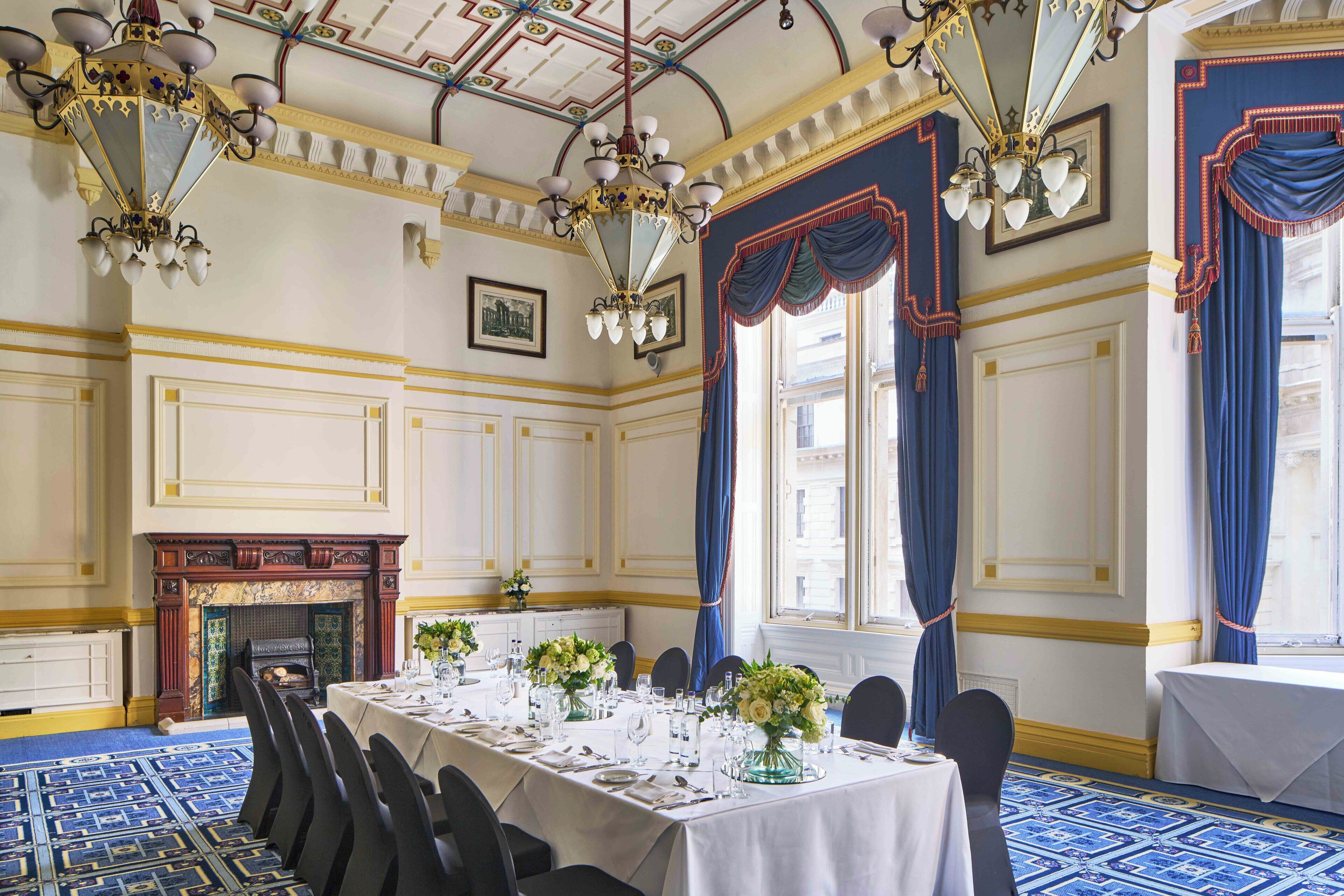 A formal dining room with a long table set for a meal, a fireplace, and large windows with blue curtains.
