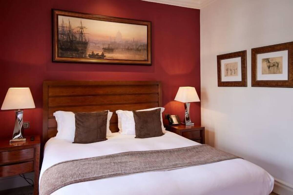 A hotel room with a large bed, two bedside tables, a headboard, a painting above the bed, and two framed pictures on the wall.
