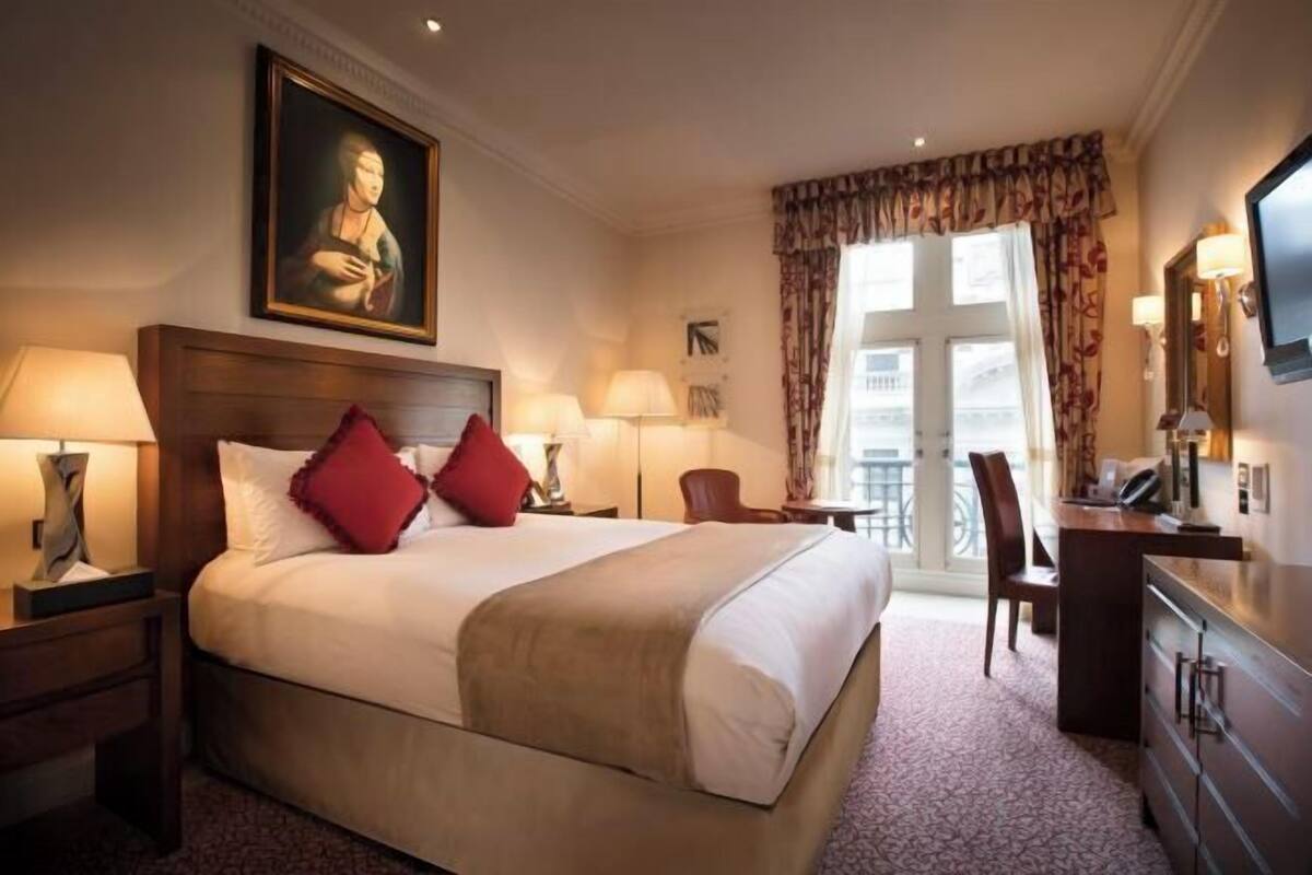 A hotel room with a large bed, two bedside tables with lamps, a desk with a chair, a television, and a large window with curtains.