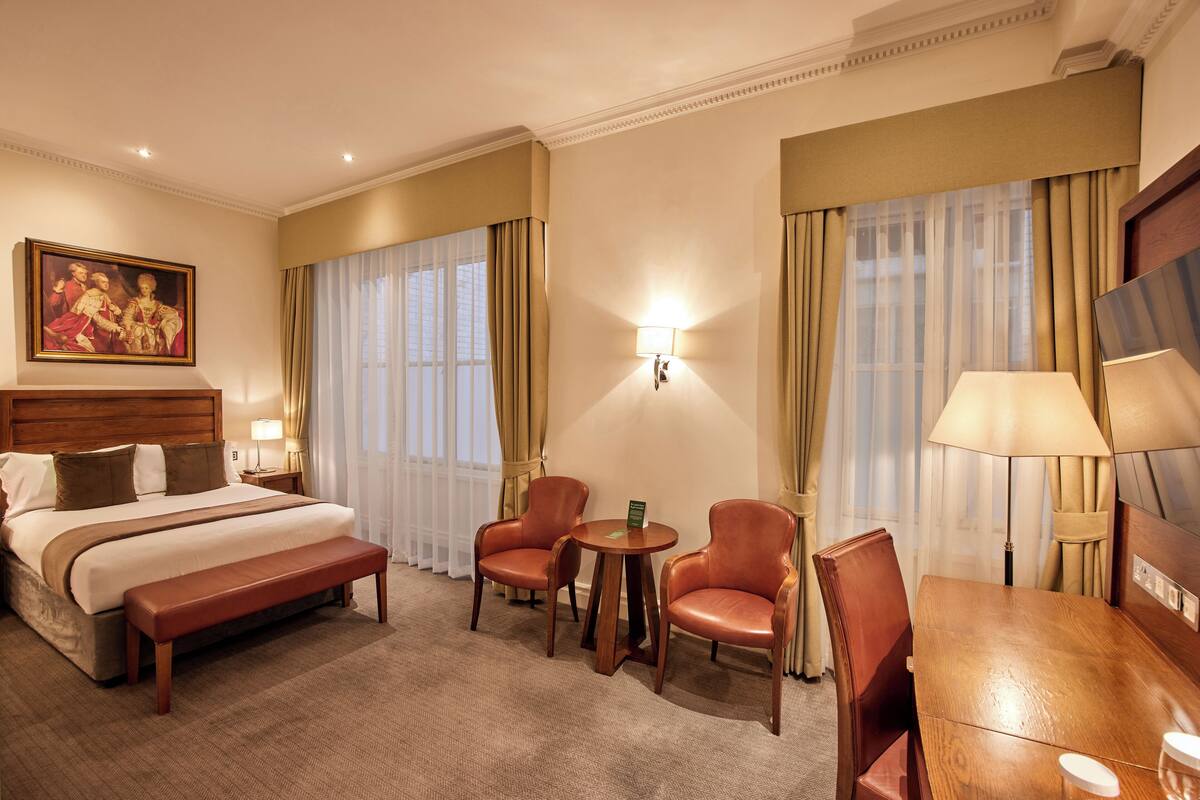 A hotel room with a large bed, a bench, two leather chairs, a small table, and a painting on the wall.