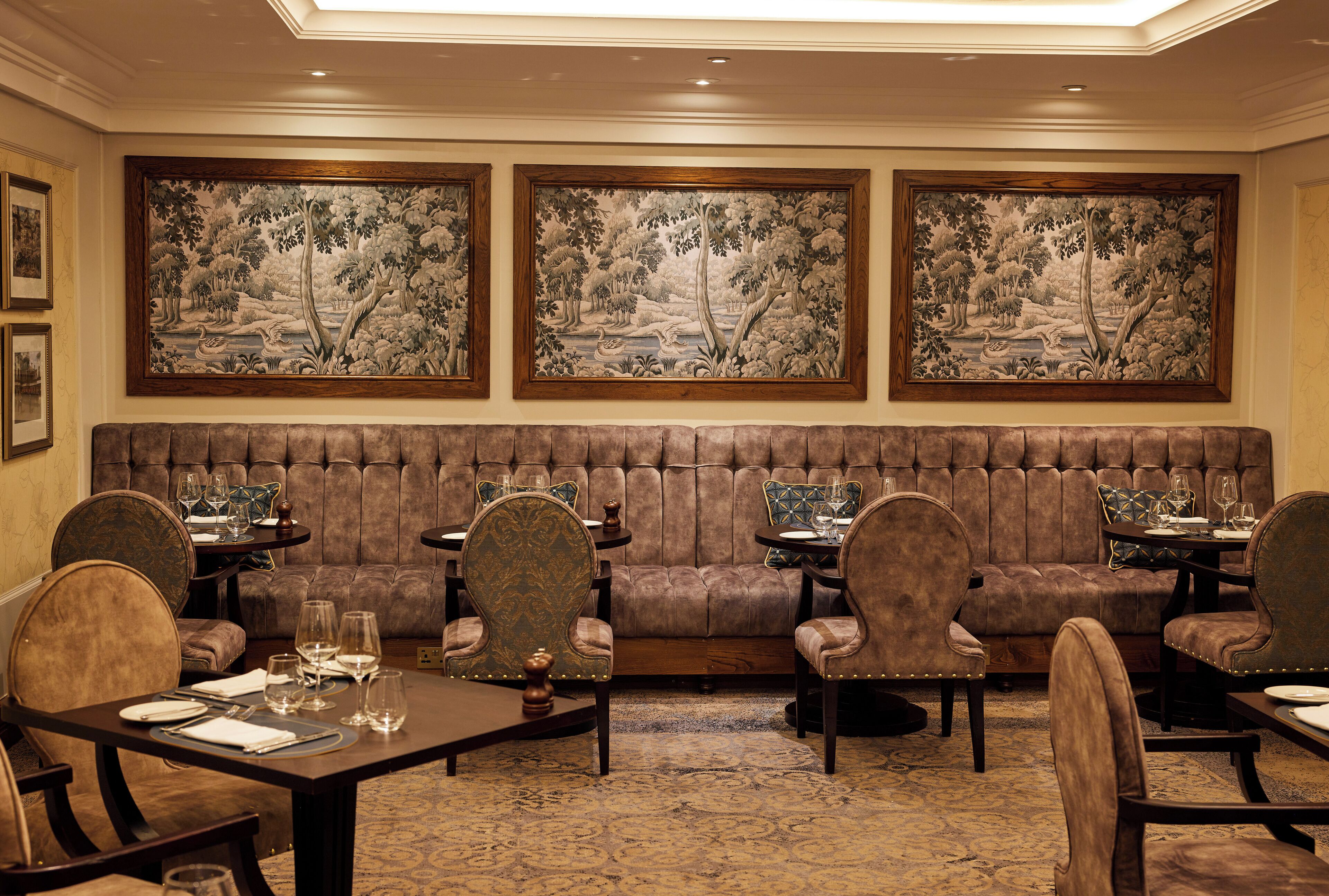 A dining area with a brown upholstered bench, multiple dining tables, and chairs. There are three large framed paintings on the wall depicting landscapes.