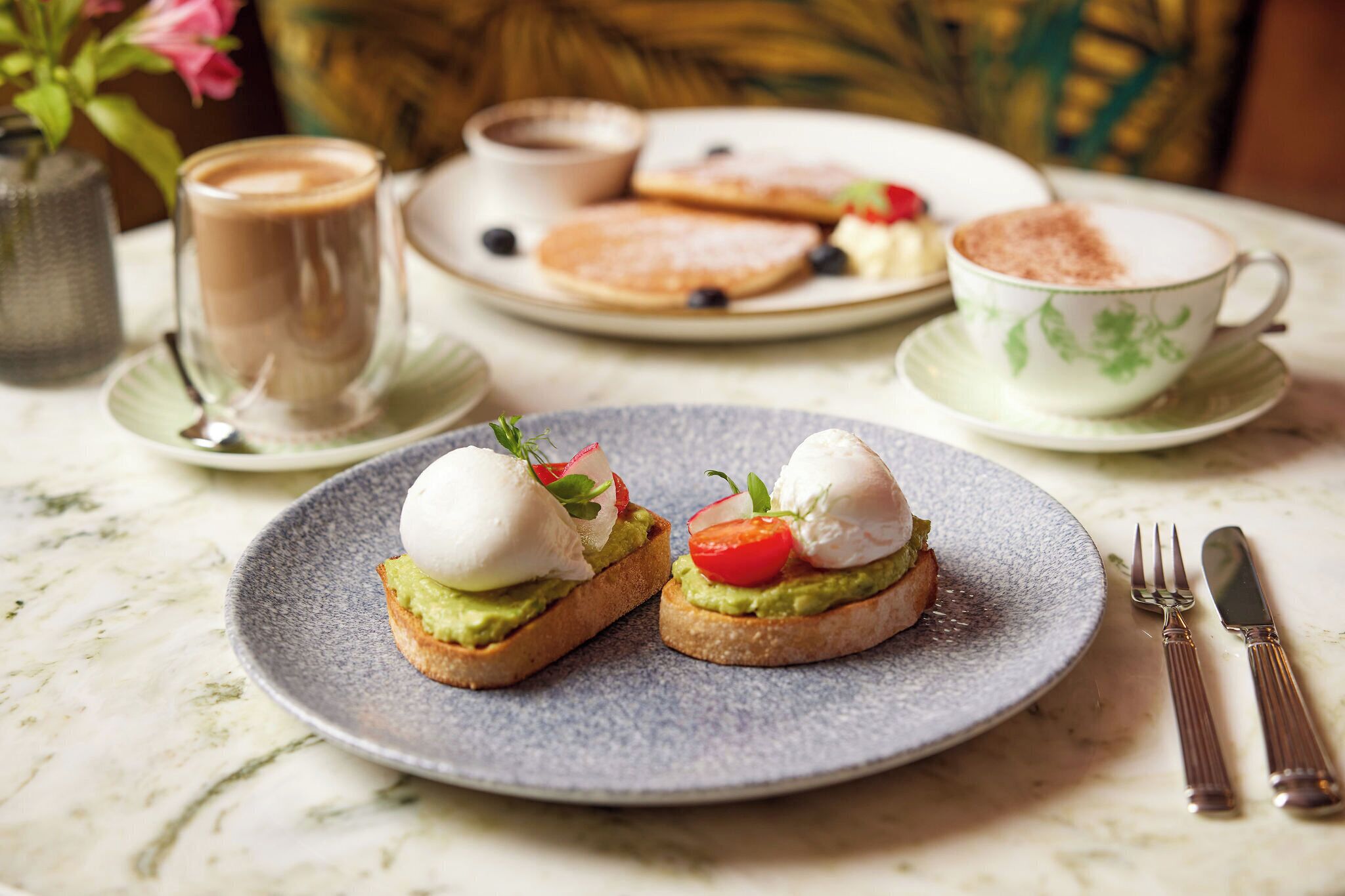 A breakfast setting with two open-faced sandwiches on a plate, a cup of coffee, and a plate with pancakes and a strawberry.