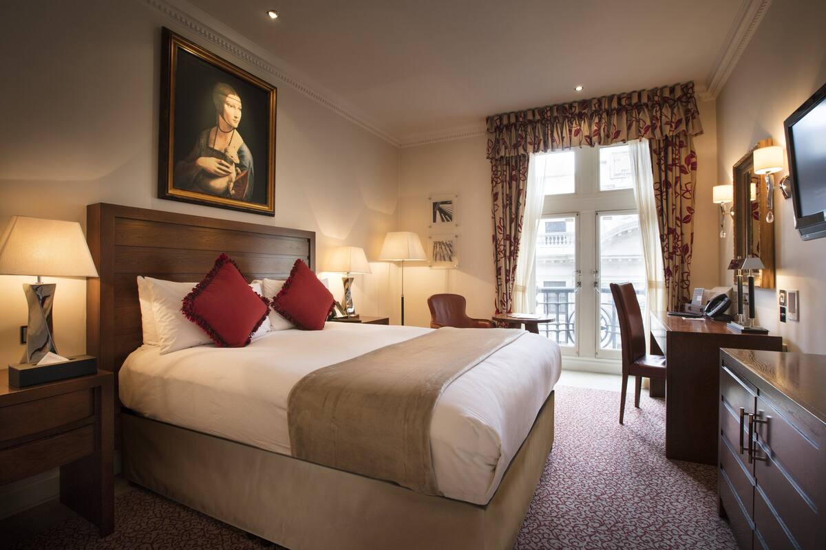 A hotel room with a large bed, two bedside lamps, a desk with a chair, a television, and a window with curtains.
