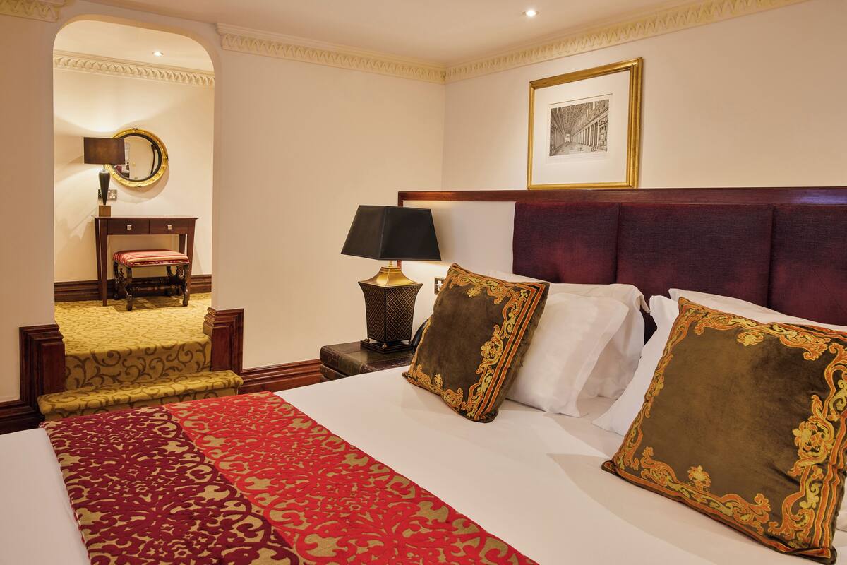 A hotel room with a large bed, a bedside table with a lamp, a chair, a mirror, and a framed picture on the wall.