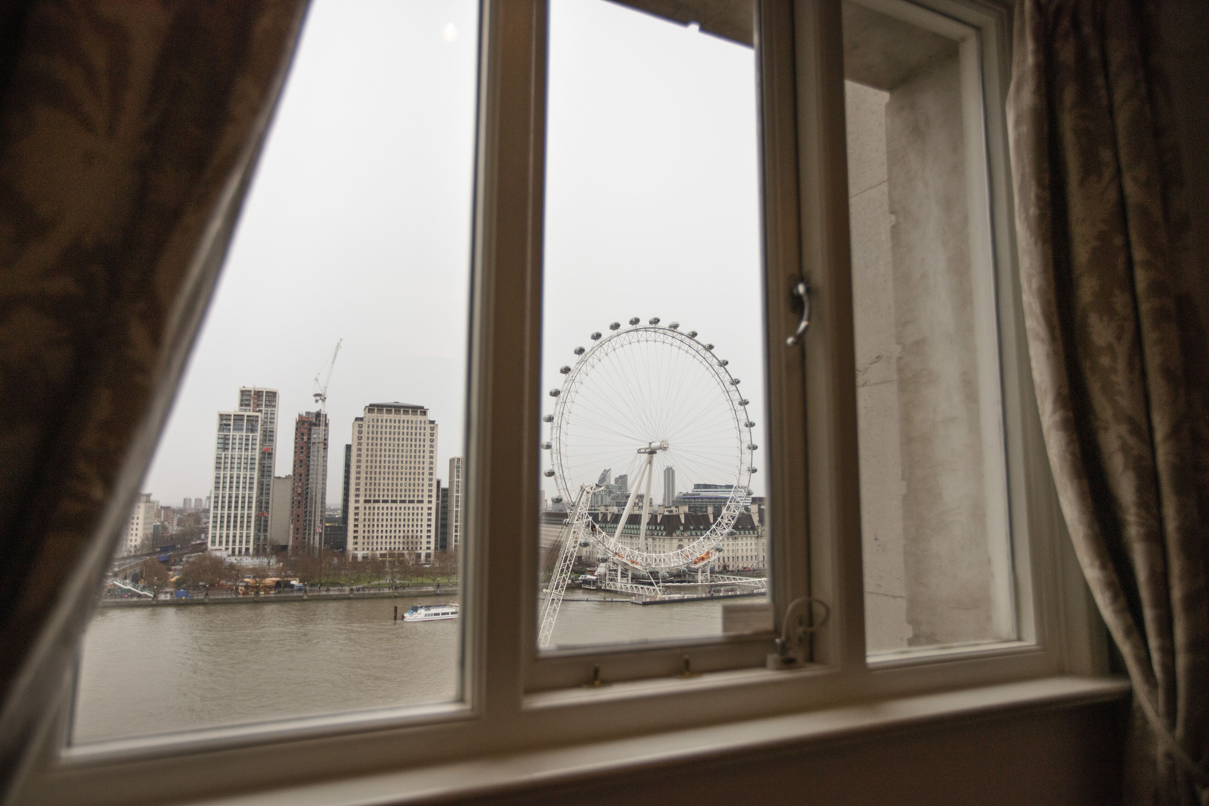 A view of a cityscape through a window with curtains, including a prominent Ferris wheel and tall buildings.