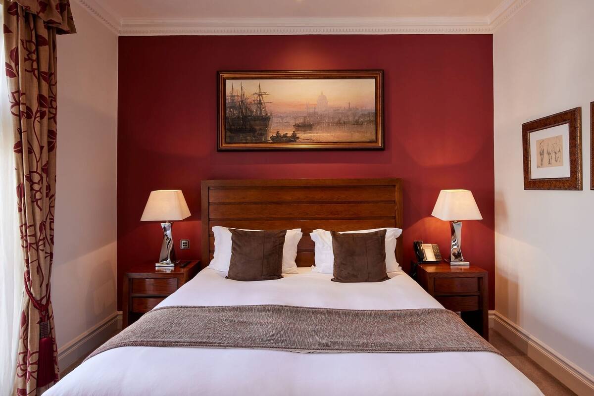 A hotel room with a large bed, two bedside tables, a painting on the wall, and a view of a cityscape.