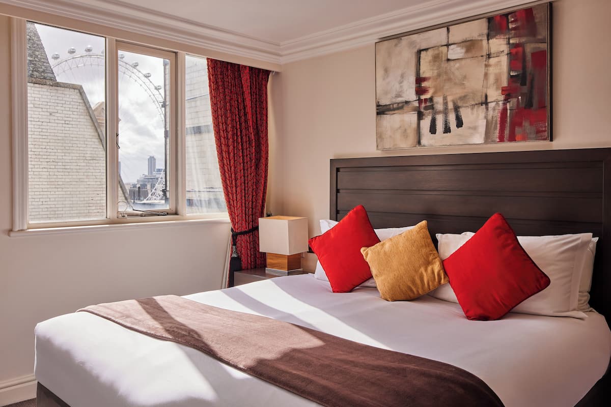 A hotel room with a large bed, a window with a view of a Ferris wheel, a nightstand with a lamp, and a painting on the wall.