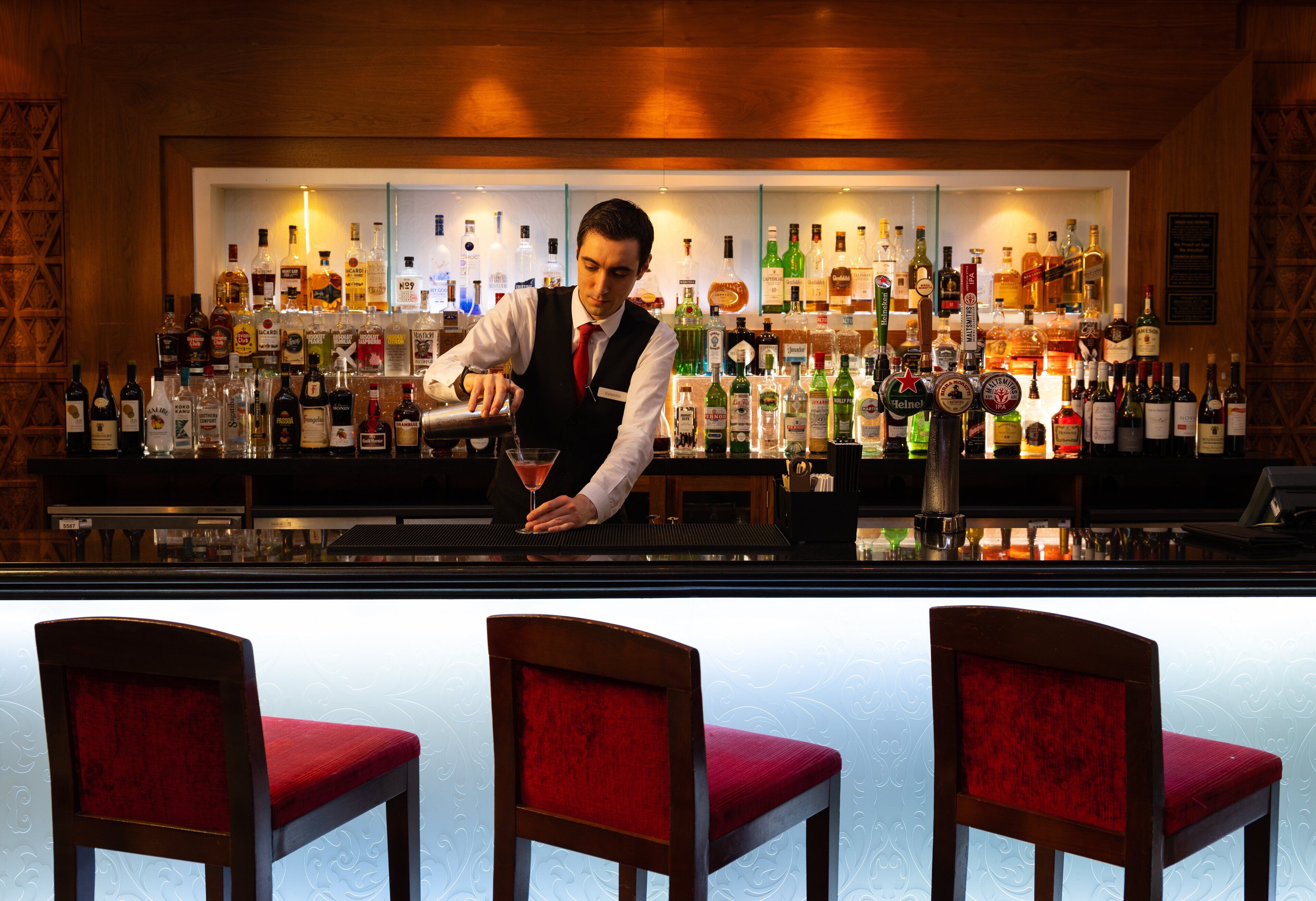 Un bar con un cantinero preparando una bebida, estantes con varias botellas de licor y tres sillas de bar tapizadas en rojo.