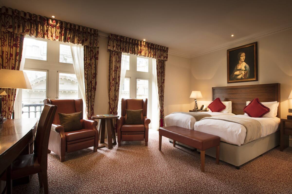 A hotel room with a bed, two chairs, a small table, a desk, and a painting on the wall.