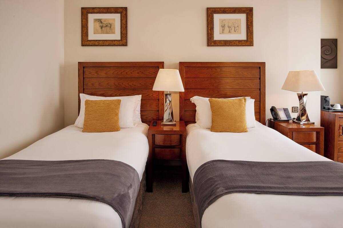 A hotel room with two single beds, wooden headboards, bedside tables with lamps, a phone, and framed artwork on the wall.