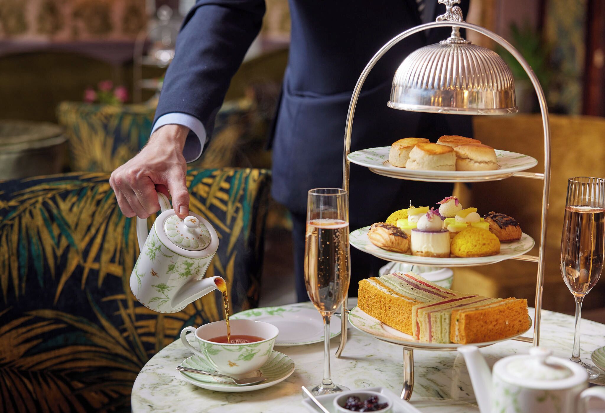 A tiered tea service with a teapot pouring into a cup, accompanied by a glass of champagne and a selection of sandwiches and pastries on a table.