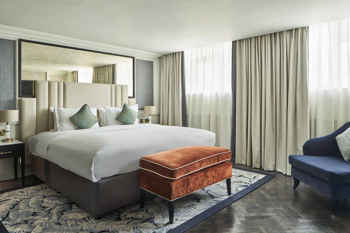 A hotel room with a large bed, a sitting area with a blue sofa, a wooden floor, and a large window with curtains.