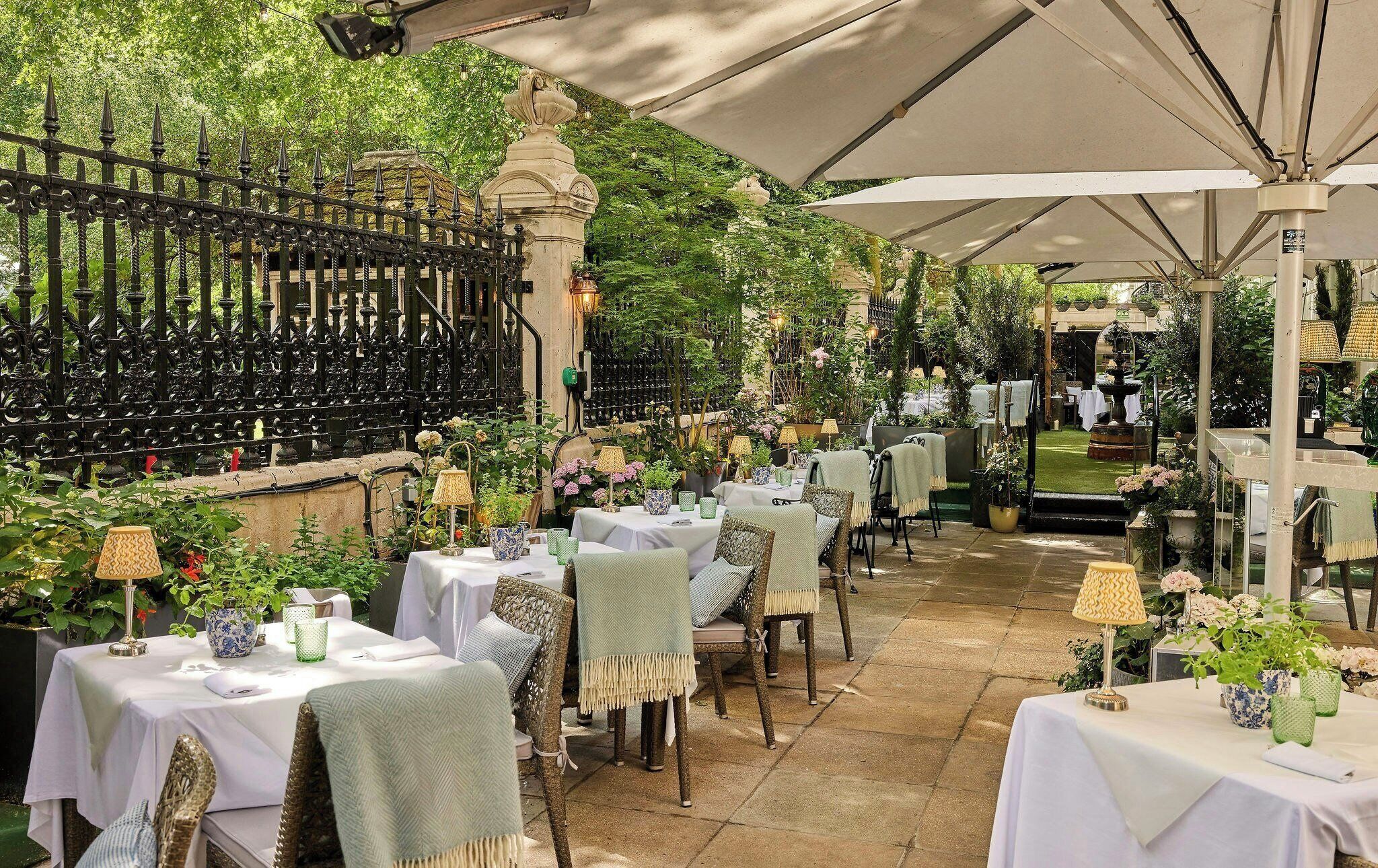 Un espacio exterior para comer con mesas y sillas, rodeado de vegetación y una valla decorativa.
