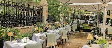 Un espacio exterior para comer con mesas y sillas, rodeado de vegetación y una valla decorativa.