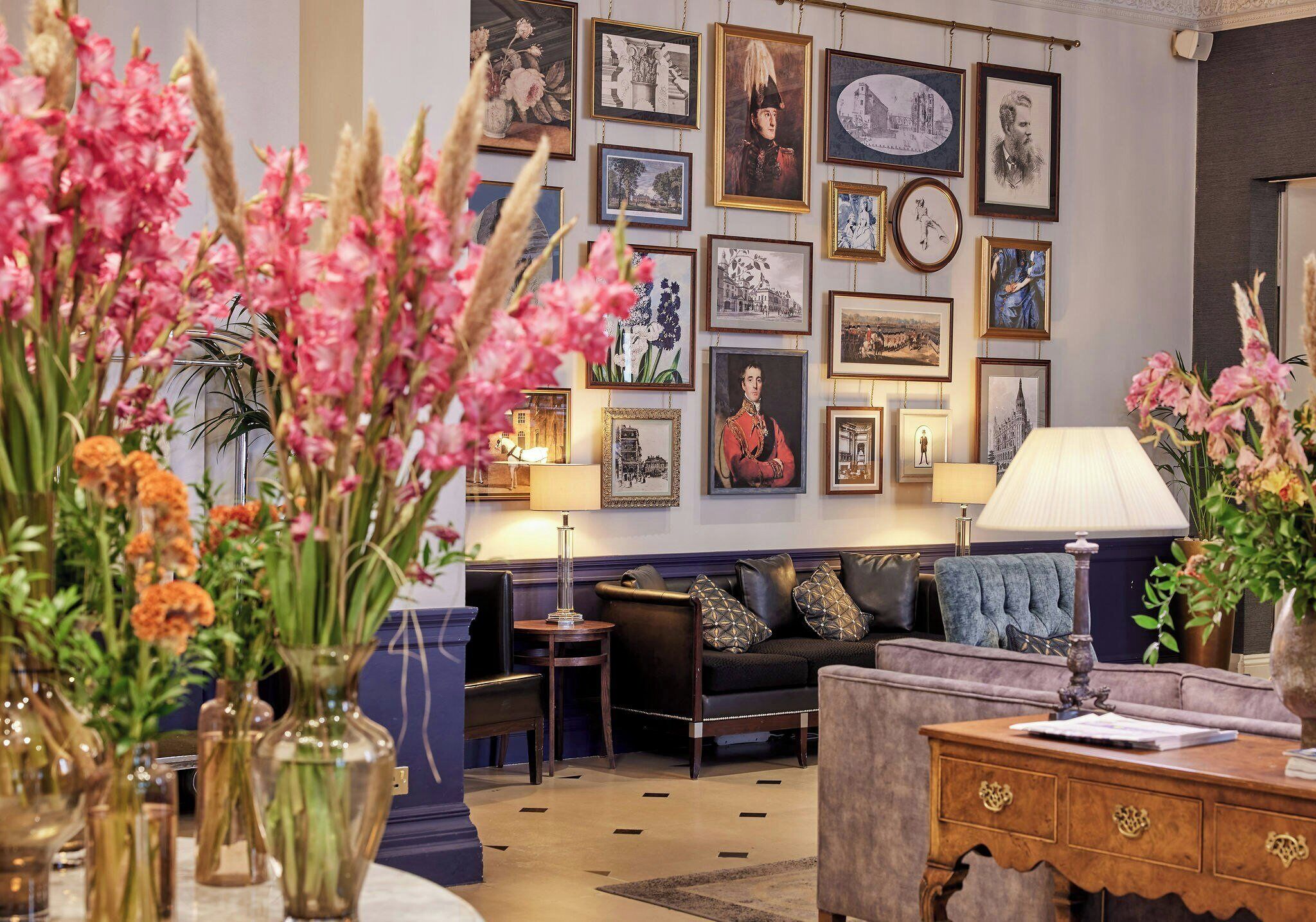 A living room with a sofa, a wooden side table, and a lamp. There are framed pictures on the wall and vases with flowers on the table.