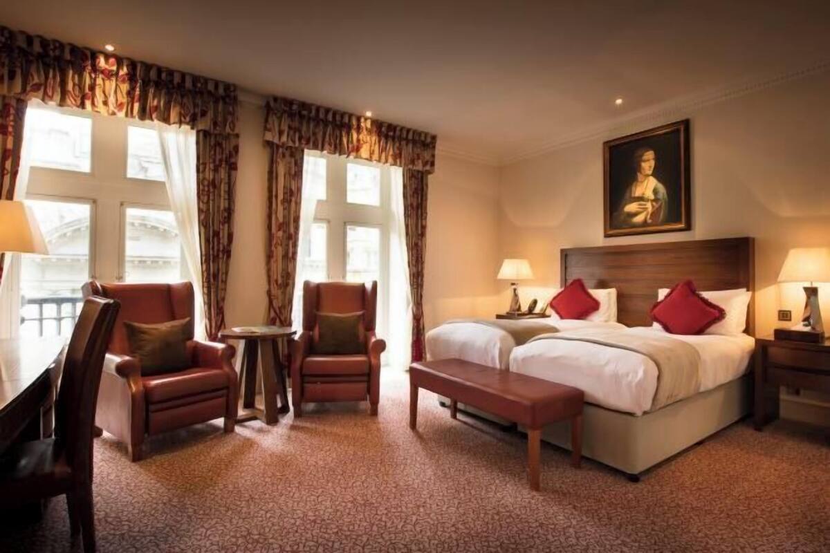 A hotel room with a bed, two chairs, a table, a bench, and a painting on the wall.