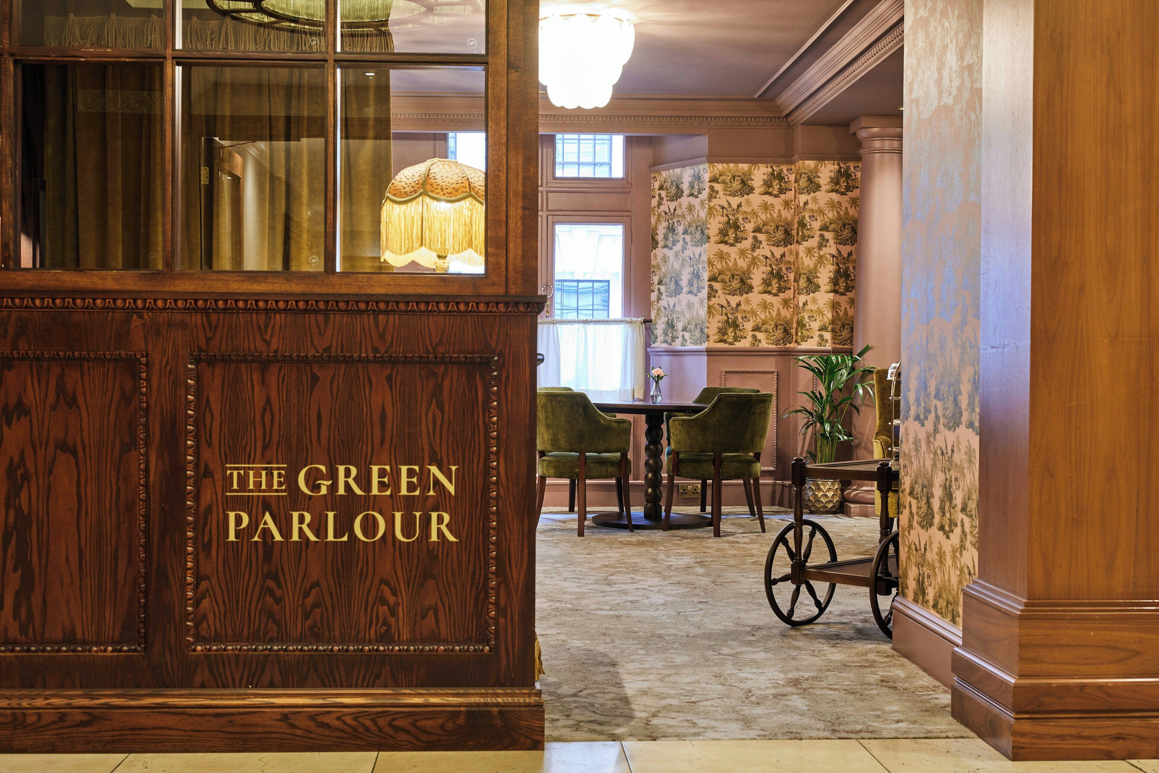 Un espace d’accueil avec un bureau en bois portant l’inscription « The Green Parlour », des chaises vertes, une petite table et une lampe décorative.