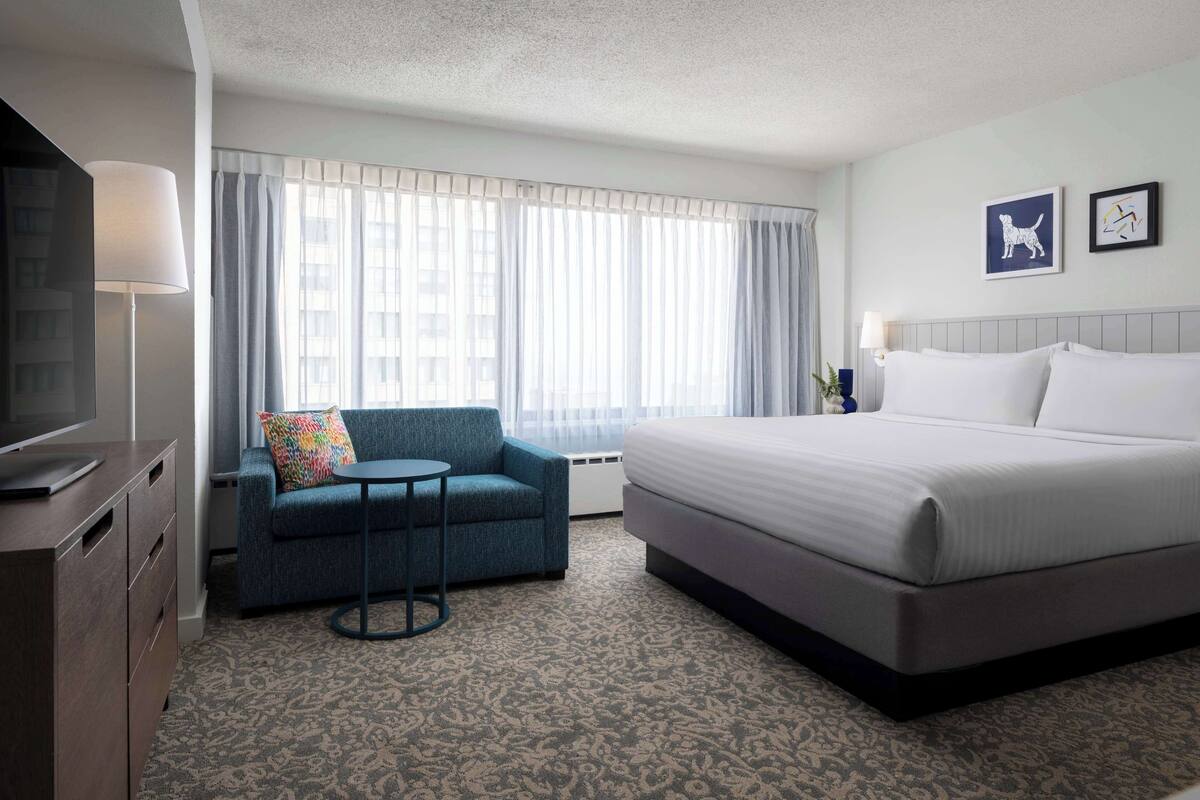 Una habitación de hotel con cama, sofá, mesita, televisor y ventana con cortinas.