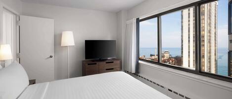 Habitación de hotel con una cama grande, un televisor sobre una cómoda de madera y vistas de la ciudad a través de un ventanal amplio.
