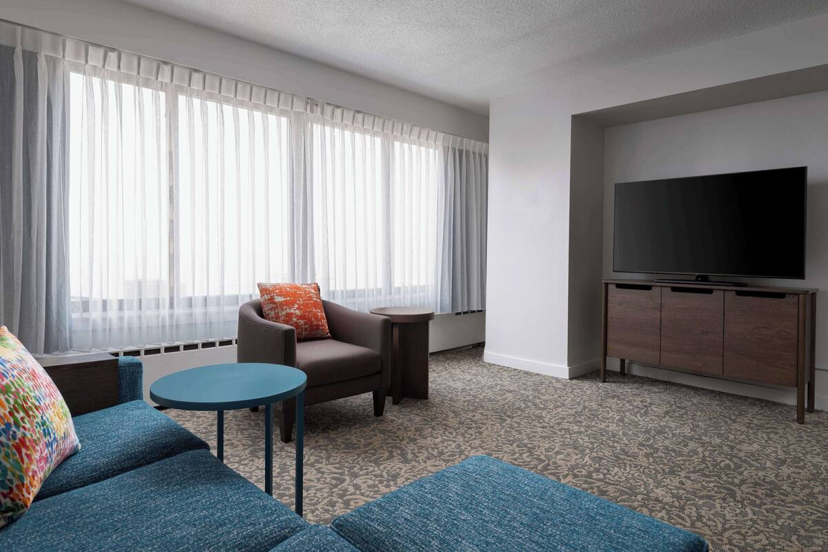 Habitación de hotel moderna con sofá, sillón, una mesita redonda y televisión sobre un mueble de madera.