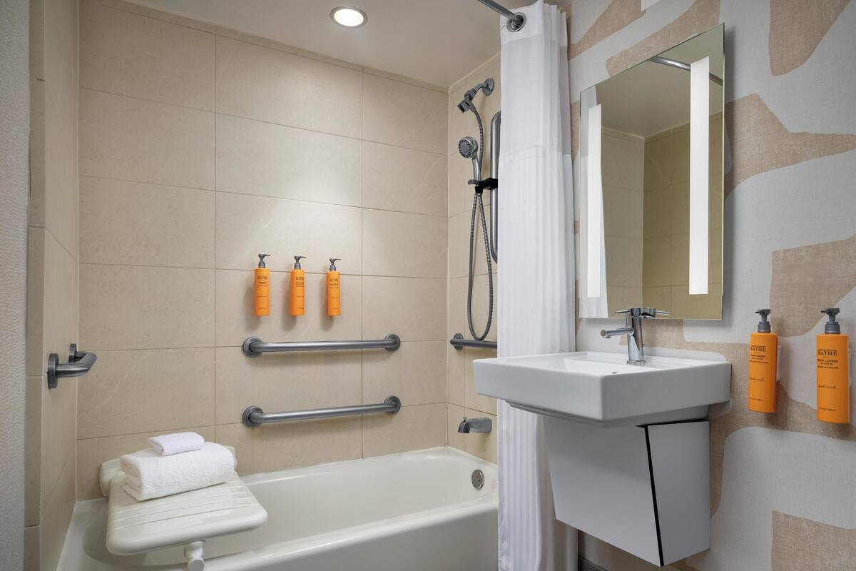 Un baño moderno con bañera, lavabo y ducha. Hay barras de apoyo, un perchero y artículos de aseo en la pared.