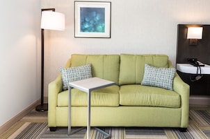 A green sofa with checkered pillows, a small table, a framed abstract painting, a floor lamp, and a bedside table with a phone and lamp.