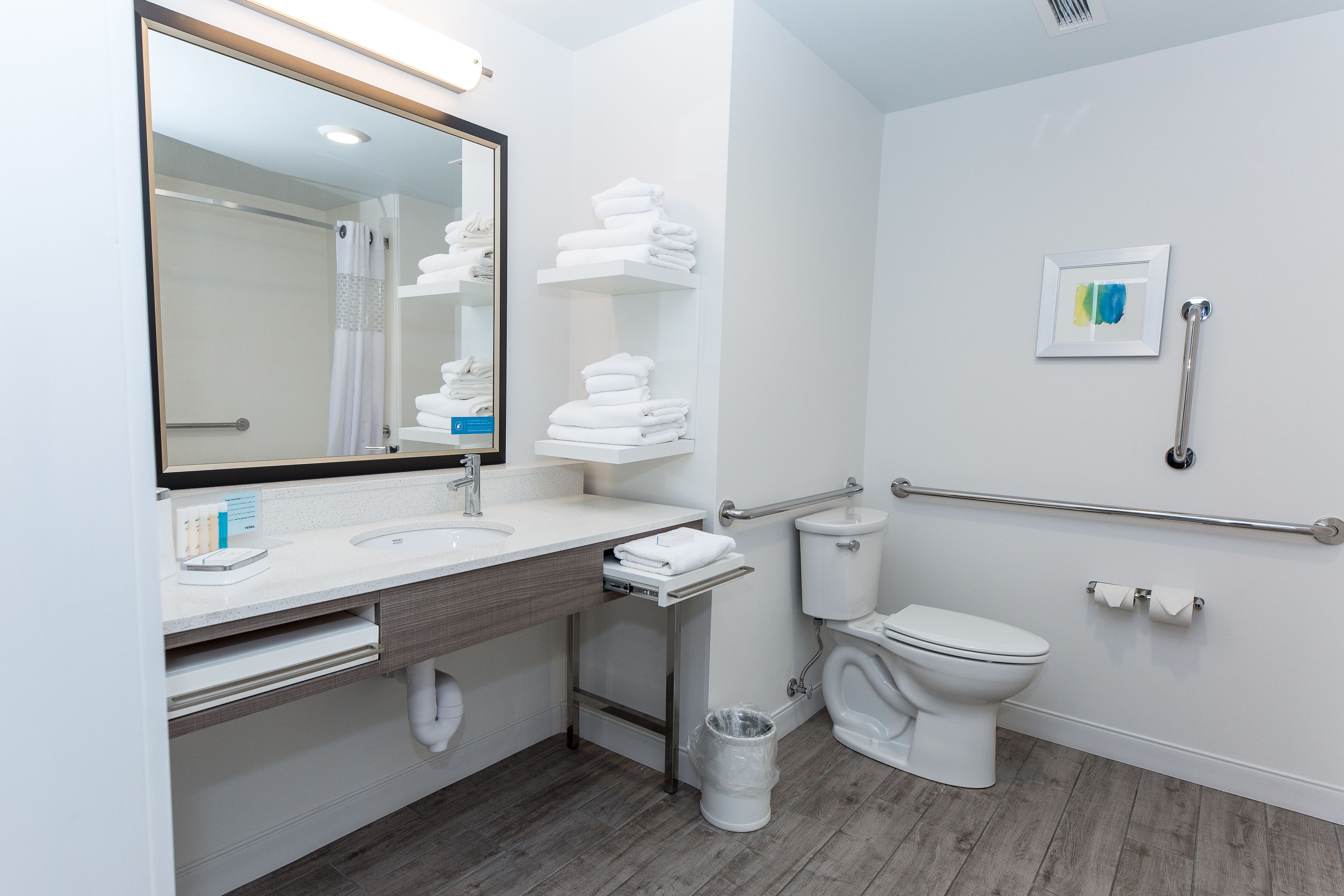 A modern bathroom with a large mirror, white sink, and toilet. There are white towels stacked on shelves, a handrail, and a trash can.