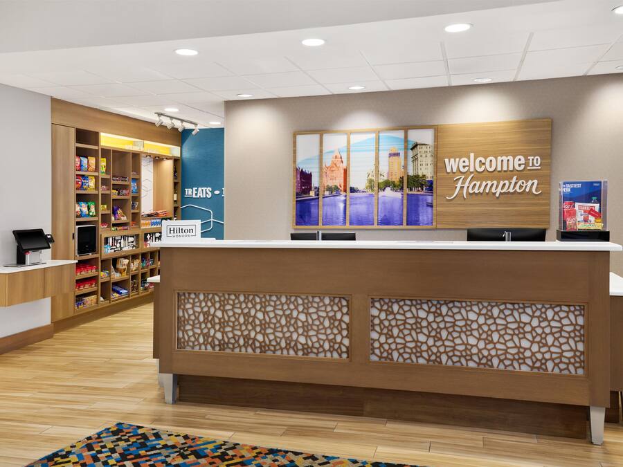 A hotel reception desk with a 'Welcome to Hampton' sign, a food display, and a digital kiosk.