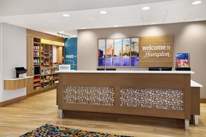 A hotel reception desk with a 'Welcome to Hampton' sign, a food display, and a digital kiosk. - Hampton Inn & Suites Syracuse North Airport Area (Liverpool)