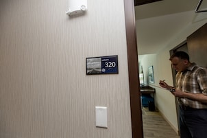 Quarto de hotel com placa indicando o número 320, um interruptor de luz e uma porta que dá para outro cômodo.
