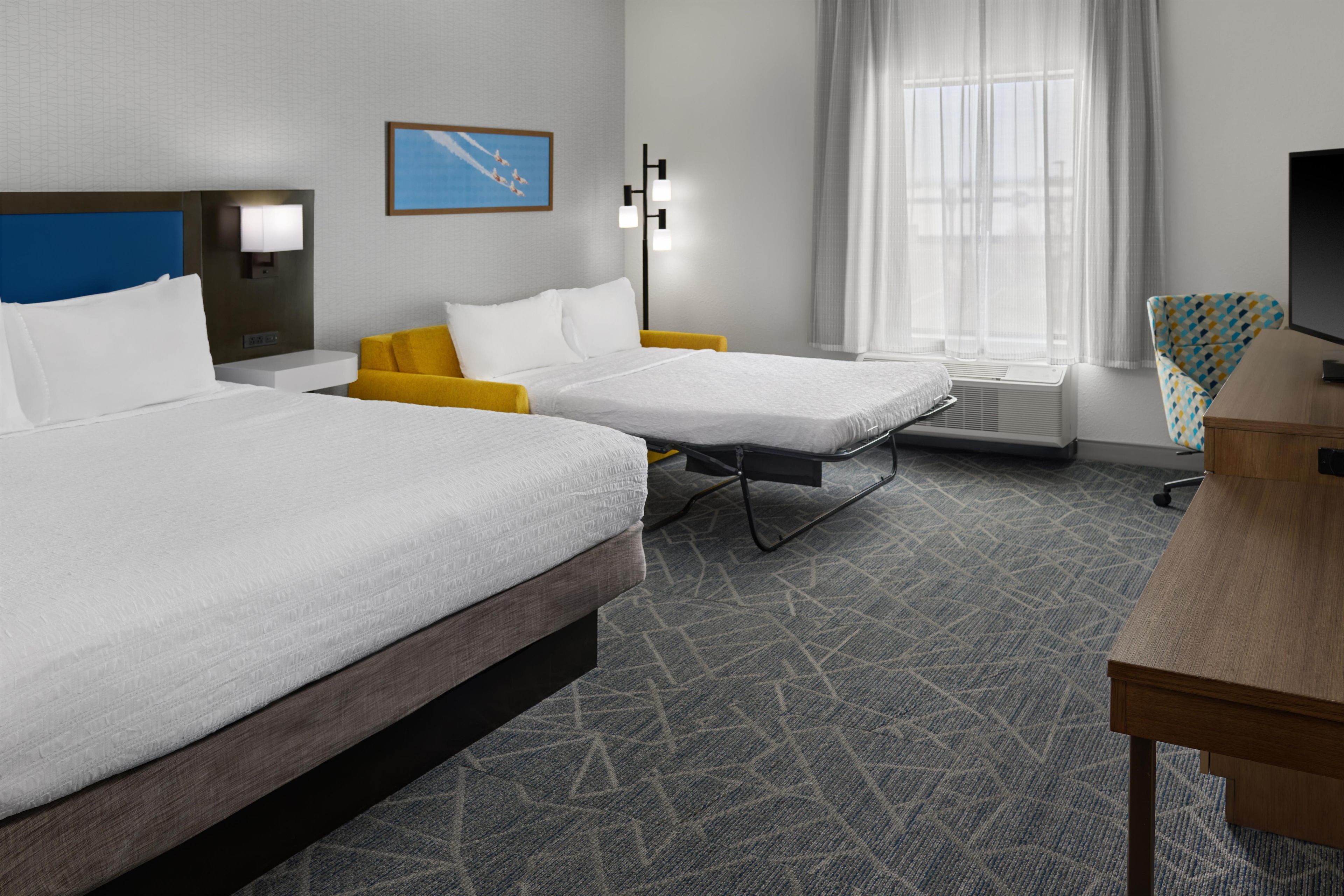 A hotel room with two beds, a desk, a chair, a TV, a window with curtains, and a wall-mounted picture.