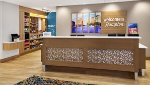 A hotel reception area with a 'Welcome to Hampton' sign, a counter, a digital screen, and a snack bar in the background. - Hampton Inn & Suites Syracuse North Airport Area (Liverpool)