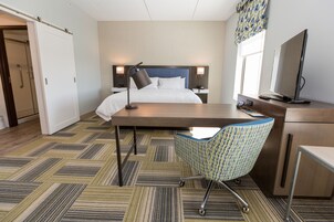 Una habitación de hotel con cama, escritorio, silla, televisor y ventana con cortinas.